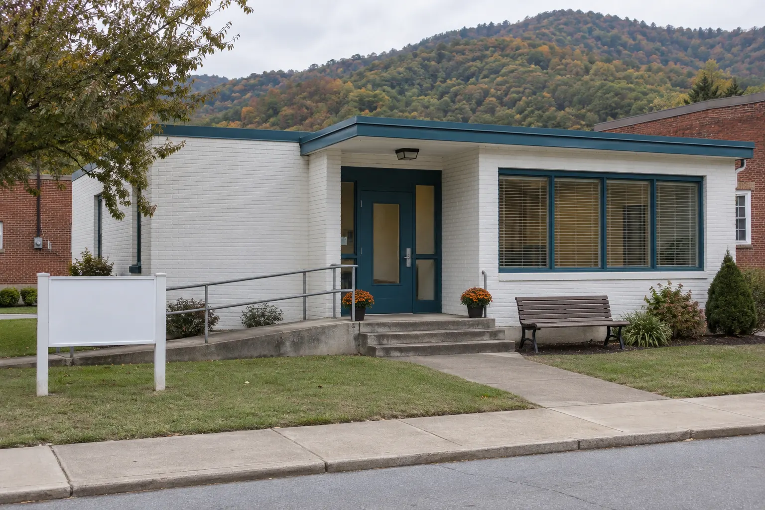 The clinic from the street: a single-storey white-brick building with a teal front door, a concrete ramp with a plain rail beside three steps, a blank sign board on the lawn, a bench, and a wooded autumn ridge behind the roof.