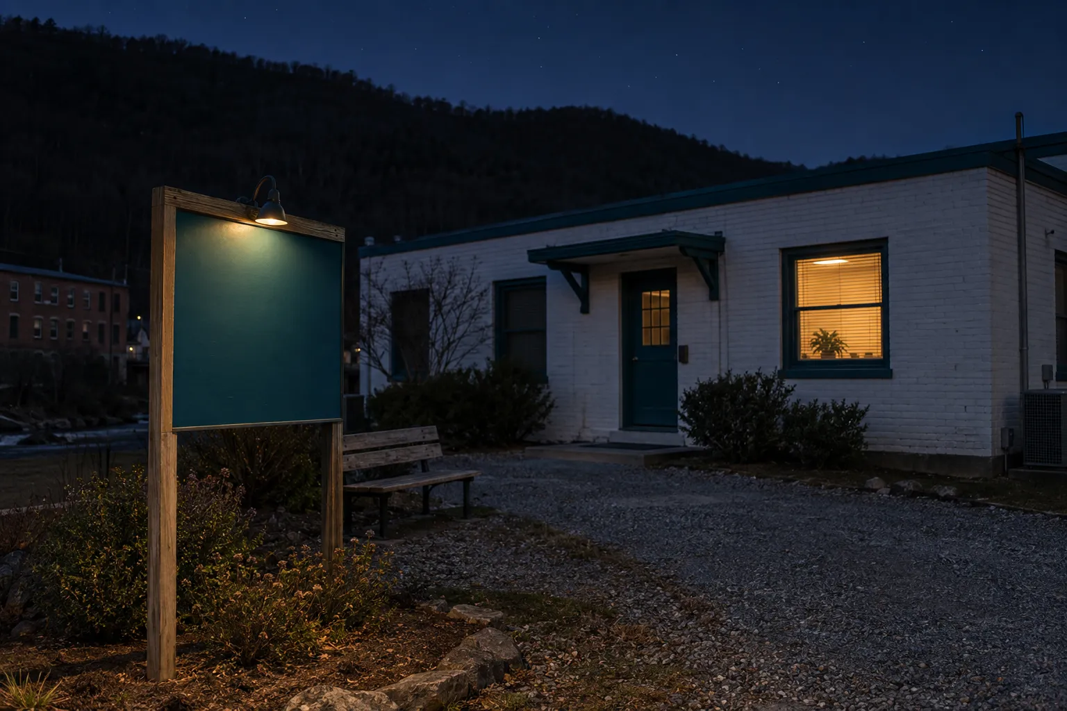 The clinic’s post-mounted sign at night: a blank teal panel lit by one small downlight, the white-brick building dark behind it with one window glowing, a gravel path, a bench and a black ridge against a deep blue sky.