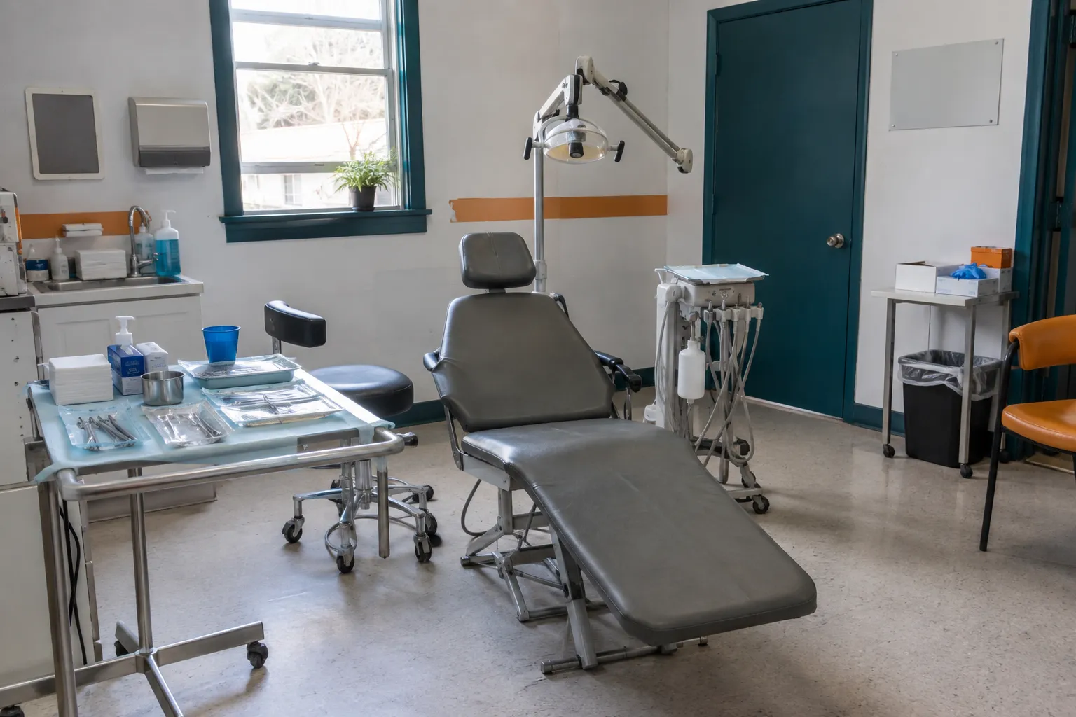 The dental-day setup before patients arrive: a portable folding dental chair, a rolling tray with sterile instrument packs in sealed pouches, a portable dental lamp, a suction unit, boxes of gloves and masks, a small window and white walls with teal trim.