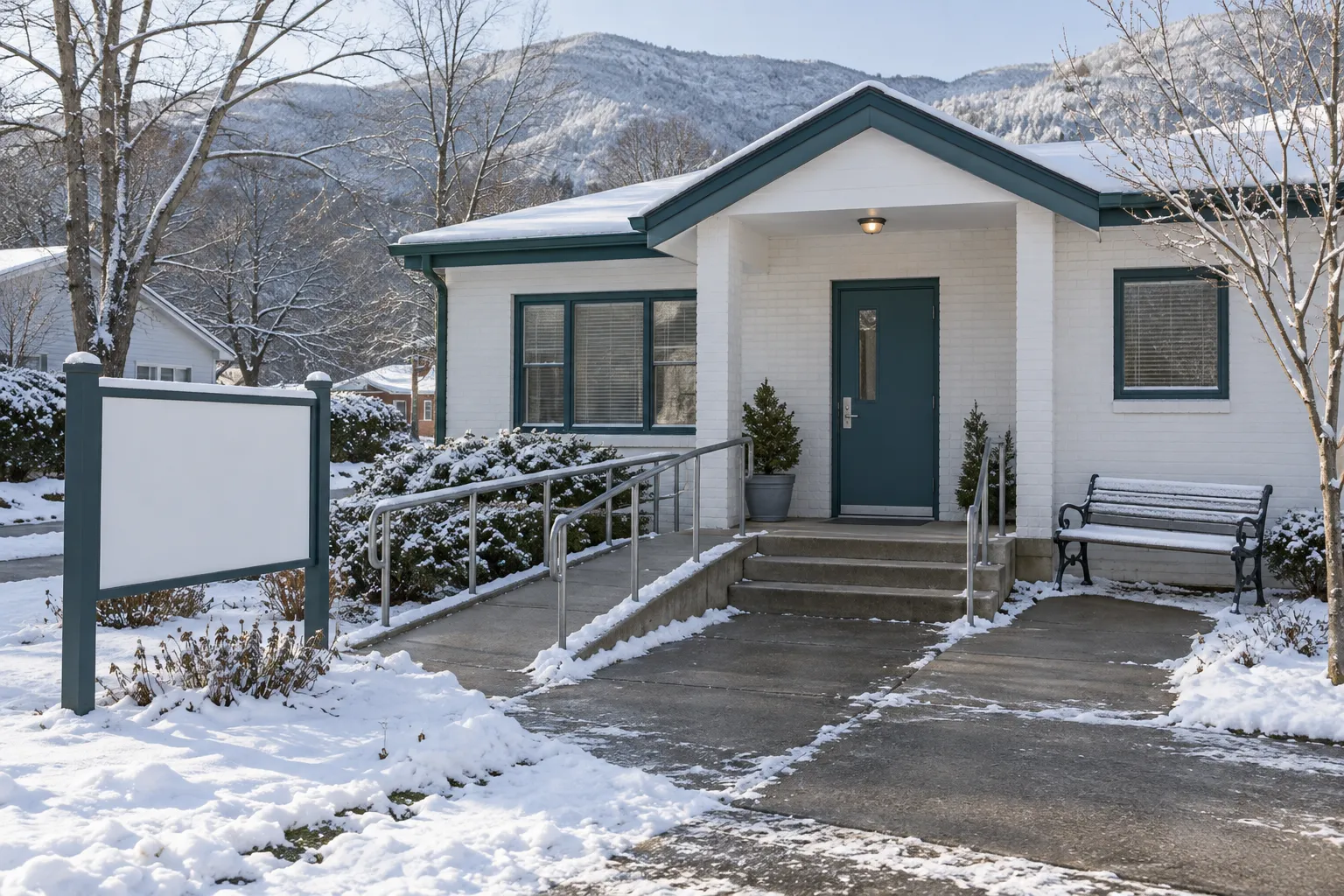 The front of the clinic on a winter morning after light snow: the teal front door, the ramp with its plain rail cleared, three steps, a bench dusted with snow, a blank sign board, bare trees and a white ridge behind under a pale low sun.
