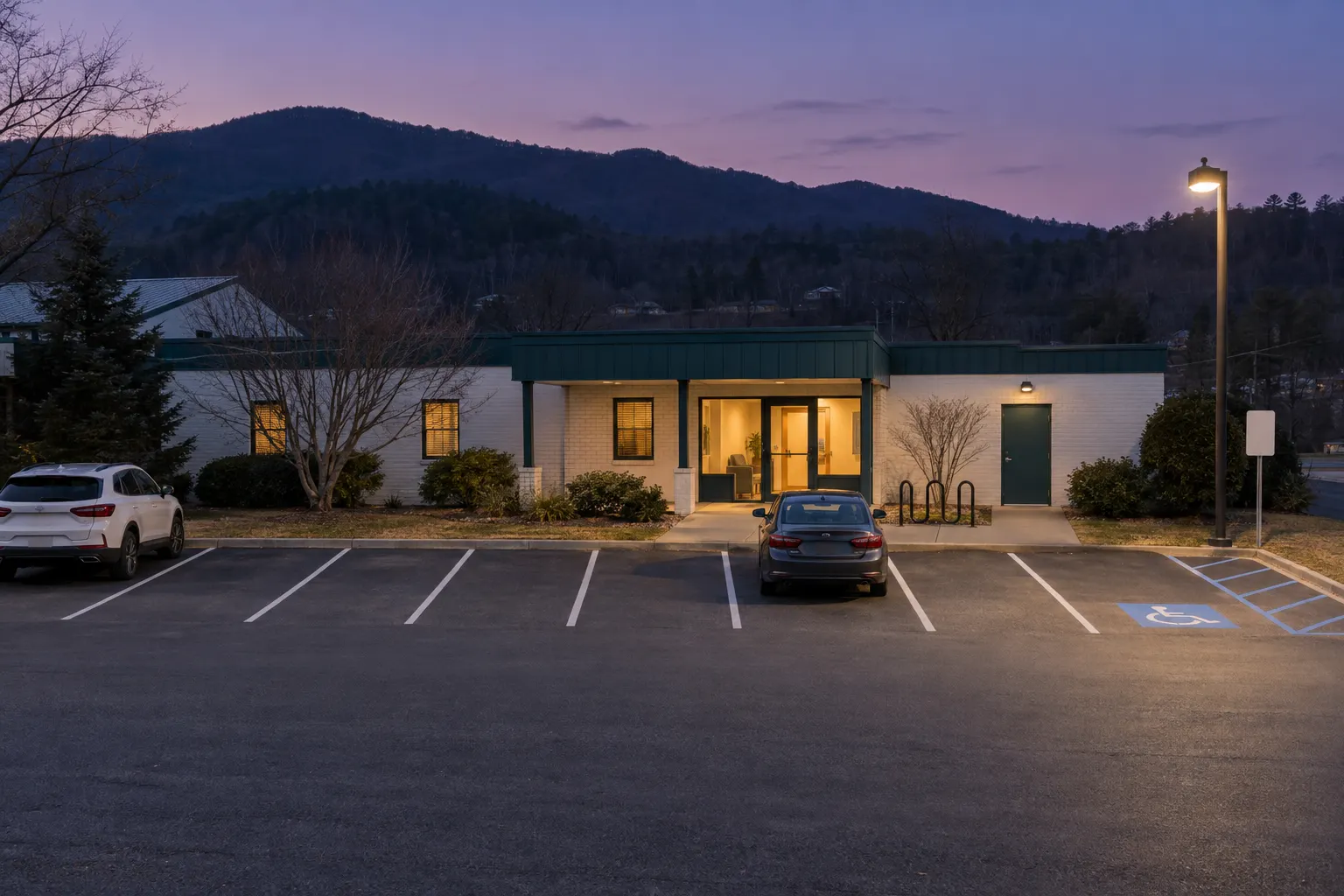 The parking lot at dusk: marked spaces, two parked cars, one lamp lit, the low white-brick clinic glowing from inside, a bike rack, an accessible parking space, and a ridge of mountains dark against a violet sky.